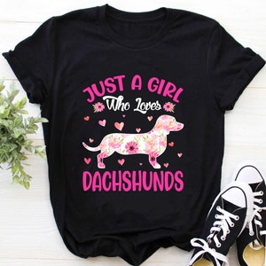 May include: Black t-shirt with pink text that reads "Just a girl who loves dachshunds". The text is surrounded by pink hearts and a floral dachshund.