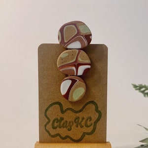 Puede incluir: Tres pendientes redondos de arcilla polimérica con un patrón abstracto marrón, blanco y verde. Los pendientes se muestran en una tarjeta marrón con el texto "Clay KC" en verde.