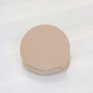 May include: A round, beige makeup sponge with a slightly textured surface.