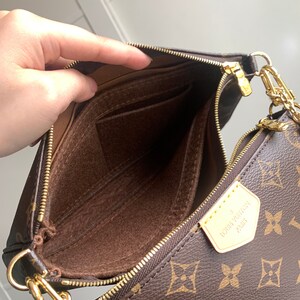May include: A brown and gold Louis Vuitton handbag with a chain strap. The bag is open and the inside is lined with brown fabric. The bag has a zipper closure and a small pocket on the inside.