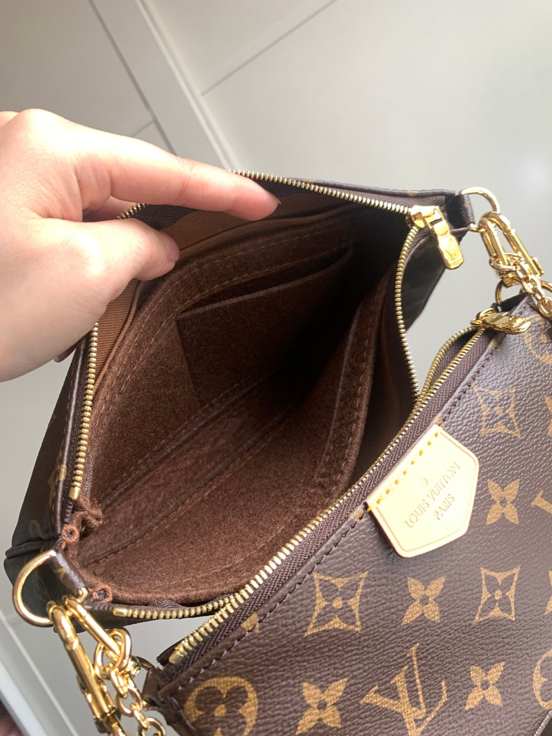 May include: A brown and gold Louis Vuitton handbag with a chain strap. The bag is open and the inside is lined with brown fabric. The bag has a zipper closure and a small pocket on the inside.