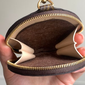 May include: A brown and beige round zippered pouch with a gold zipper and a gold chain attached. The pouch is open and the inside is lined with brown fabric.