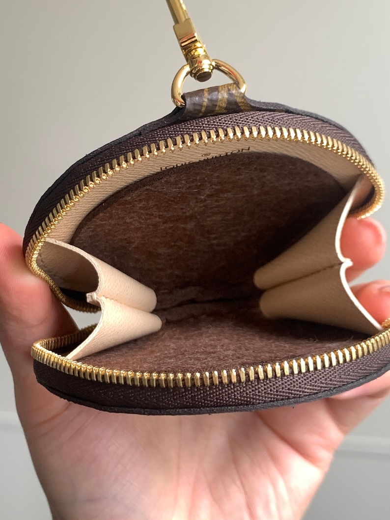 May include: A brown and beige round zippered pouch with a gold zipper and a gold chain attached. The pouch is open and the inside is lined with brown fabric.