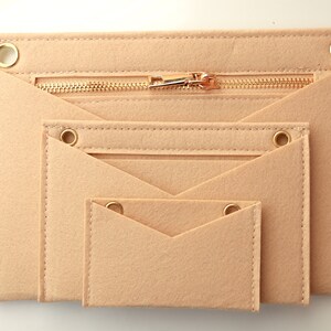 Può includere: Tre pochette in feltro beige con cerniere e occhielli dorati. Le pochette sono annidate l'una dentro l'altra, con la pochette più grande in basso e la più piccola in alto.