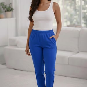 Pode incluir: Uma mulher vestindo uma regata branca e calças azuis. As calças têm um design de perna reta e bolsos. A mulher está sorrindo e em pé em um quarto bem iluminado.