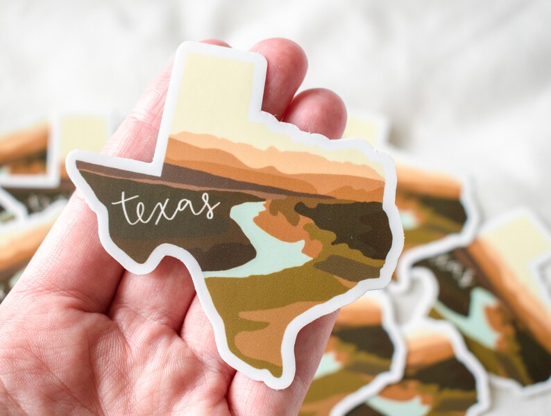 Puede incluir: Una pegatina con la forma del estado de Texas, con un dise&ntilde;o de paisaje en tonos marrones, beige y verdes. La palabra "Texas" est&aacute; escrita en cursiva en la pegatina.