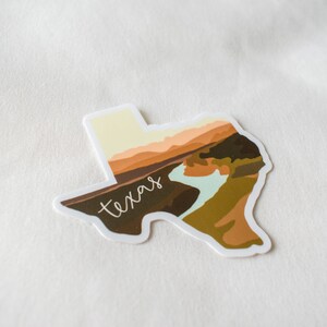 Puede incluir: Una pegatina con la forma de Texas, con un dise&ntilde;o de paisaje en tonos marrones, beige y azul claro. La palabra "texas" est&aacute; escrita en cursiva en la imagen.