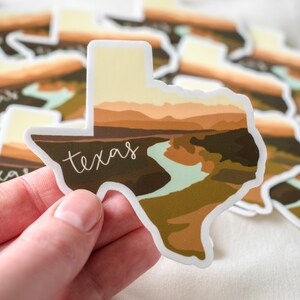Puede incluir: Una colecci&oacute;n de pegatinas con forma de Texas. Cada pegatina presenta un dise&ntilde;o de paisaje con un r&iacute;o y monta&ntilde;as en tonos marrones, beige y verdes. La palabra "texas" est&aacute; escrita en cursiva en cada pegatina.