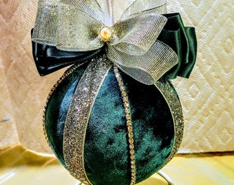 May include: A green velvet Christmas bauble with silver glitter and rhinestones. The bauble is decorated with a silver and black bow and is sitting on a white stand.
