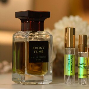 May include: Four glass bottles of perfume with gold caps. The largest bottle is labeled "Ebony Fume Eau de Parfum 50ml". The smaller bottles are labeled "Rebottled by Aromasample" and "Name Brand".
