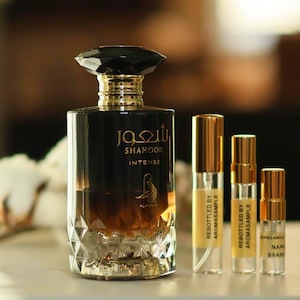 May include: A black and gold glass bottle of Shahoor Intense perfume with a black diamond-shaped stopper. The bottle is sitting on a table with three smaller bottles of perfume in front of it. The smaller bottles are labeled "Rebottled by Aromasample".