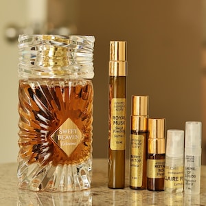 Puede incluir: Una colecci&oacute;n de frascos y viales de perfume. Una botella grande y ornamentada de vidrio con una tapa dorada y una etiqueta que dice "Sweet Heaven Tobacco" est&aacute; junto a varios viales m&aacute;s peque&ntilde;os con tapas y etiquetas doradas. Las botellas est&aacute;n sobre una superficie de m&aacute;rmol.