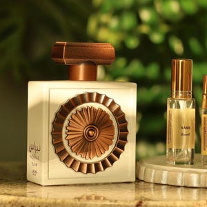 May include: An off-white perfume bottle with a bronze-colored cap and decorative front, alongside several smaller sample bottles. The samples are displayed on a round, off-white tray. The background features greenery and a window with horizontal blinds.
