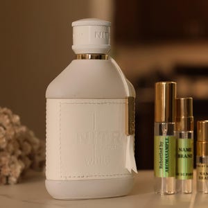 May include: A white bottle of perfume with a gold cap and a white label. The bottle is sitting on a table with three smaller bottles of perfume. The smaller bottles are clear glass with gold caps and labels. The labels on the smaller bottles have text that reads "Personally by Aromasample" and "NAME BRAND".