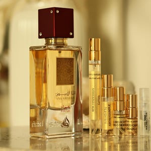 K&ouml;nnte beinhalten: Ein Parf&uuml;mflakon mit weinrotem Verschluss und goldenen Akzenten, beschriftet mit "ANA ABIYEDH ROUGE" auf Arabisch und Englisch. Mehrere kleinere Probenflaschen sind davor angeordnet, mit goldenen und transparenten Fl&auml;schchen. Der Hintergrund ist unscharf.