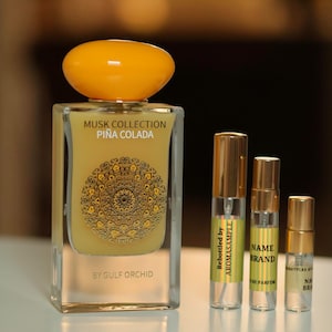 May include: Three glass bottles of perfume with gold labels. The largest bottle is yellow and has the text "Musk Collection Piña Colada by Gulf Orchid". The smaller bottles are clear and have labels with the text "Rebottled by Aromasample", "Name Brand", and "Bottled by Name Brand".