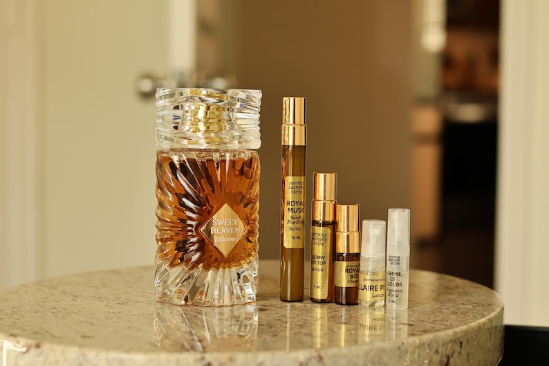 Puede incluir: Una colecci&oacute;n de frascos y viales de perfume. Una botella grande y ornamentada de vidrio con una tapa dorada y una etiqueta que dice "Sweet Heaven Tobacco" est&aacute; junto a varios viales m&aacute;s peque&ntilde;os con tapas y etiquetas doradas. Las botellas est&aacute;n sobre una superficie de m&aacute;rmol.