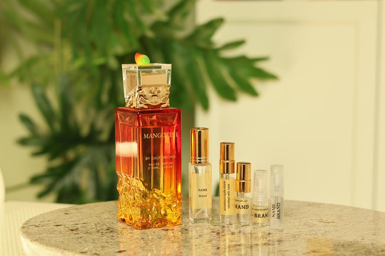 May include: A perfume bottle with a square, orange-to-yellow gradient design, topped with a decorative cap. Several smaller sample bottles and vials are arranged in front of the perfume bottle. The text "MANGO" is visible on the perfume bottle.