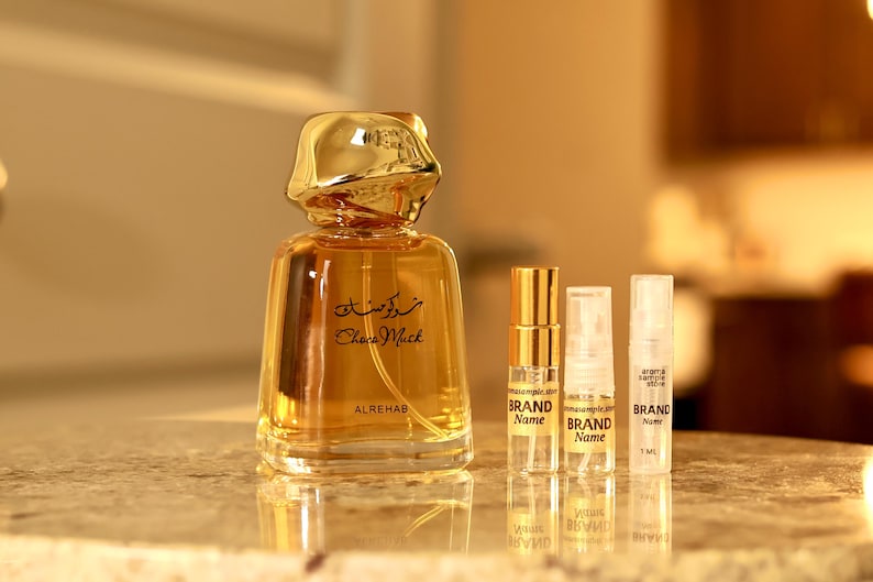May include: A perfume bottle with a gold-colored cap and a clear glass body, containing a golden liquid. The bottle has Arabic script and the words "Choco Musk" printed on it. There are also three smaller sample bottles.