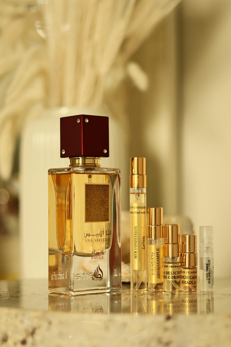K&ouml;nnte beinhalten: Ein Parf&uuml;mflakon mit weinrotem Verschluss und goldenen Akzenten, beschriftet mit "ANA ABIYEDH ROUGE" auf Arabisch und Englisch. Mehrere kleinere Probenflaschen sind davor angeordnet, mit goldenen und transparenten Fl&auml;schchen. Der Hintergrund ist unscharf.