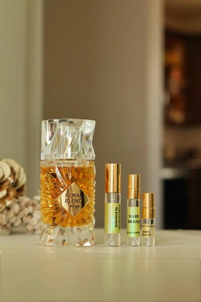 May include: Three perfume bottles. The largest bottle is clear glass with a gold label that reads 'Royal Blend Vintage'. The two smaller bottles are clear glass with gold caps and labels that read 'Rebottled by Aromasample' and 'NAME BRAND Eau de Parfum'.