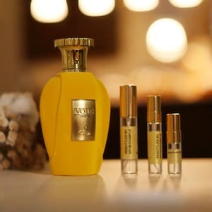 May include: A yellow perfume bottle with a gold cap and label, next to three smaller sample bottles. The label on the large bottle reads "Voux Zeste". The sample bottles are labeled "Rebottled by AromaSample".