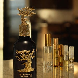 May include: A dark perfume bottle with a gold tree design and a matching tree-shaped cap. The bottle is labeled "Eternal Vanille" by Lattafa Pride. Several smaller perfume samples are arranged beside it.