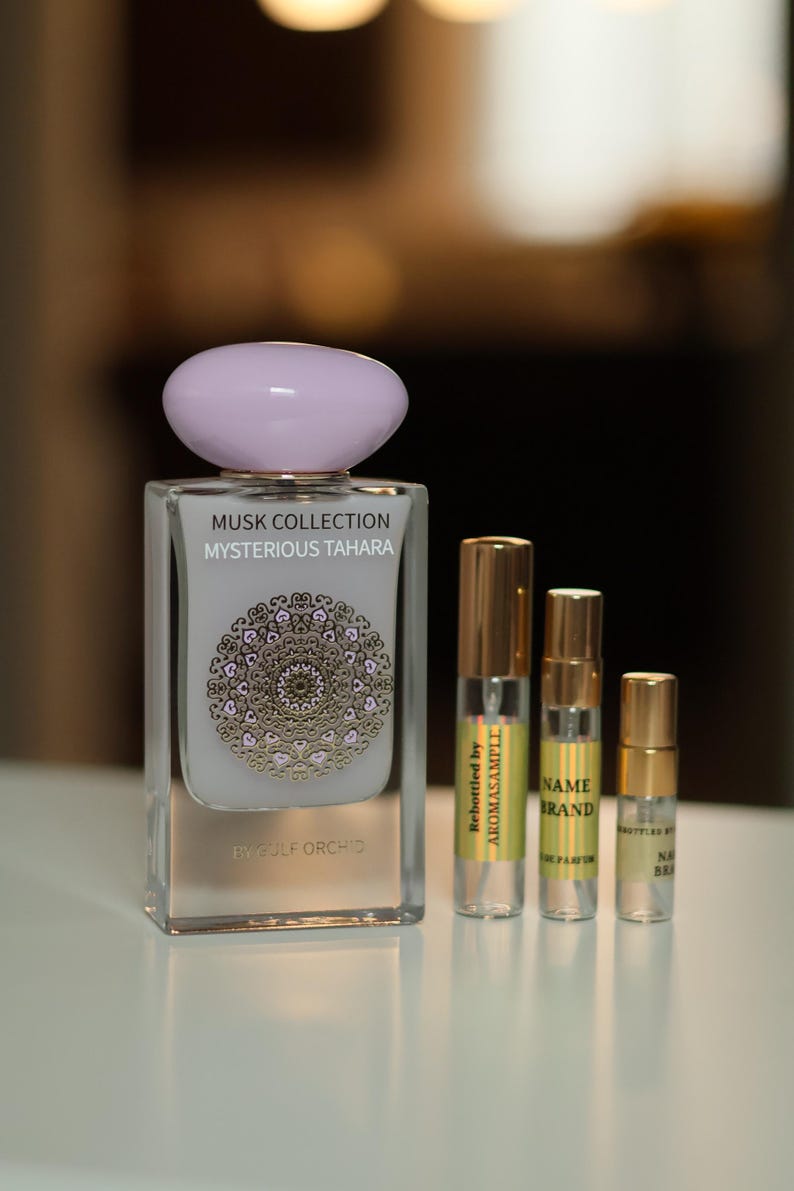 May include: A clear glass bottle of perfume with a light purple lid. The bottle is labeled 'MUSK COLLECTION MYSTERIOUS TAHARA BY GULF ORCHID'. Three smaller bottles of perfume are also visible, each with a gold cap and a label. The labels on the smaller bottles read 'Rebottled by AROMASAMPLE', 'NAME BRAND', and 'BOTTLER BY NAME BRAND'.