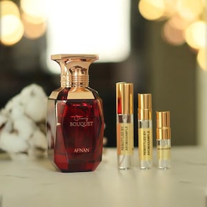 May include: A red glass bottle of Cherry Bouquet perfume by Afnan. The bottle has a gold cap and a gold label with the brand name and fragrance name. There are three smaller bottles of perfume in the background.