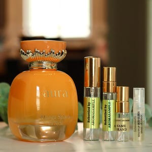 May include: A clear glass bottle with a gold lid and a label that says "aura" and "Mango Splash Extreeme". The bottle is sitting on a table with four smaller bottles next to it. The smaller bottles are labeled "Rebottled by AROMASAMPLE".