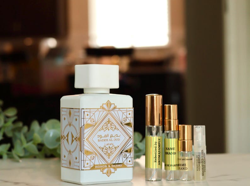 Puede incluir: Una botella blanca de perfume Bader Al Oud con detalles dorados y el texto "بديع المود" y "BADER AL OUD" en la parte delantera. La botella est&aacute; sobre una mesa con cuatro botellas de perfume m&aacute;s peque&ntilde;as delante.