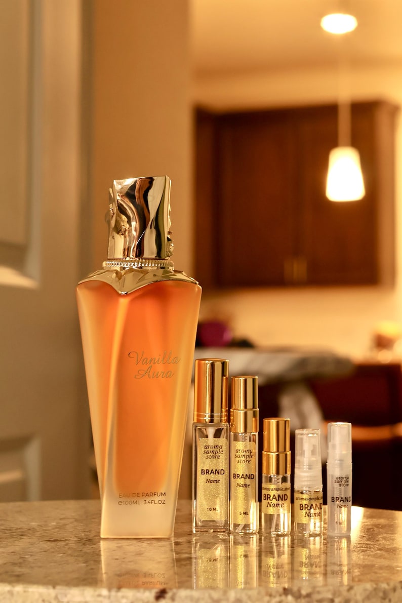 以下が含まれることがあります： 「Vanilla Aura」の文字が入った琥珀色の香水瓶と、いくつかのサンプルバイアルが展示されています。大きなボトルには「EAU DE PARFUM e100ml 3.4FLOZ」と記載されています。小さなバイアルには金色のキャップが付いており、「aroma sample store」と「BRAND Name」と記載されています。