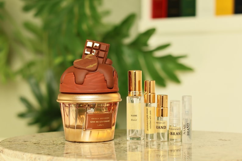 K&ouml;nnte beinhalten: Eine goldfarbene Parf&uuml;mflasche in Cupcake-Form mit schokoladen&uuml;berzogenem Deckel, gekr&ouml;nt von einem Schokoriegel und einem Herz. Daneben stehen mehrere Parf&uuml;mflaschen mit goldenen Kappen. Der Text auf der Cupcake-Flasche lautet "Choco Intense Give Me Gourmand".
