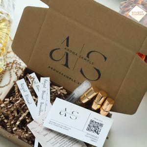 以下が含まれることがあります： 「AROMA SAMPLE」の文字が入った開いた段ボール箱には、香水サンプル、名刺、香りのストリップが入っています。「ROYAL BLEND」と書かれたものを含む、いくつかの香水ボトルが見えます。