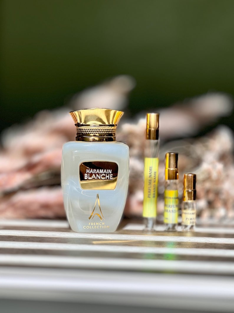K&ouml;nnte beinhalten: Eine durchsichtige Glasflasche mit Harmain Blanche-Parfum, goldener Kappe und Etikett. Die Flasche steht auf einer Holzoberfl&auml;che mit zwei kleineren Parf&uuml;mflaschen im Hintergrund.