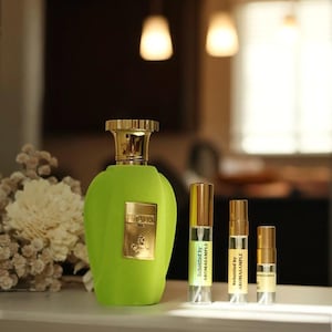 May include: A green glass bottle of perfume with a gold cap and label. The bottle is sitting on a white surface with three smaller bottles of perfume. The smaller bottles are labeled "Rebottled by Aromasample".