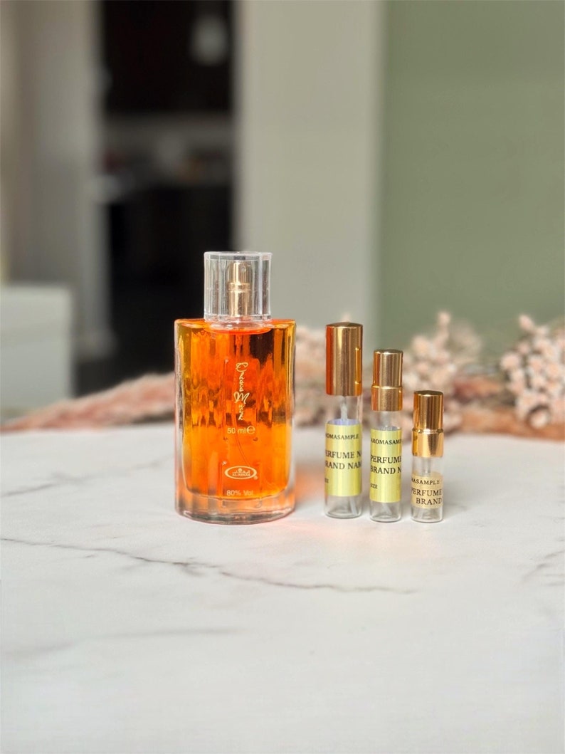 May include: Amber-colored perfume bottles in various sizes. A larger 50ml bottle with a clear glass body and gold cap is displayed next to three smaller perfume sample bottles with gold caps. The smaller bottles have labels indicating 'PERFUME SAMPLE' and 'BRAND NAME'. The larger bottle is labeled '50 ml e' and has a small label indicating '80%'. The overall theme is perfume and fragrance samples.