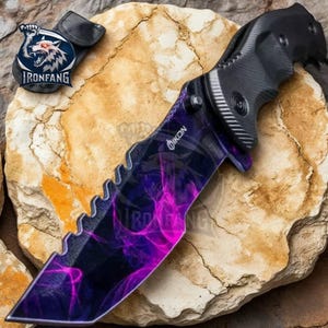 May include: A black folding knife with a purple and black patterned blade. The knife has a serrated edge and a black handle. The brand name "OIKON" is printed on the blade.