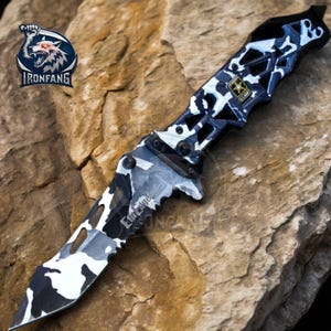 May include: A black and white camouflage folding knife with a serrated blade and a black handle. The handle has a star design and the text "Elite Force" is printed on the blade.