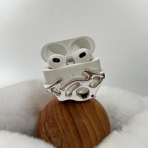May include: White wireless earbuds in a white case with a silver, abstract, decorative design. The case is sitting on a wooden ball.