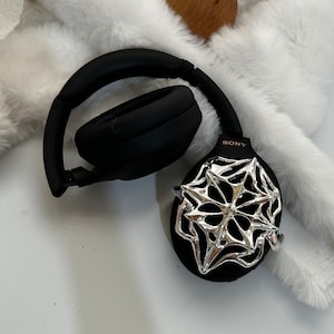 May include: Black over-ear headphones with a silver decorative accent. The headphones are resting on a white, fluffy surface. The brand name "SONY" is visible on the side of the headphones. A wooden sphere is also in the image.