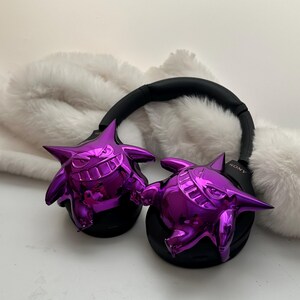 May include: Black Sony headphones with shiny purple Gengar-shaped ear covers. The headphones rest on a white, fluffy surface. The Sony logo is visible on the right earcup.