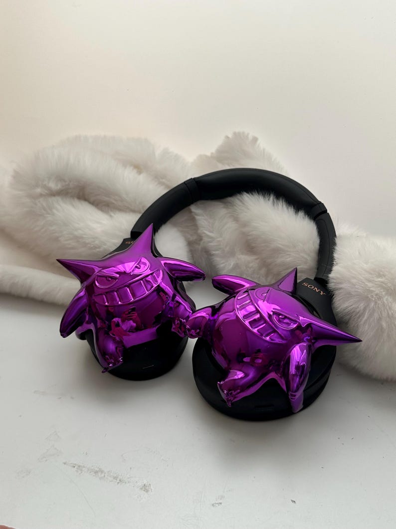 May include: Black Sony headphones with shiny purple Gengar-shaped ear covers. The headphones rest on a white, fluffy surface. The Sony logo is visible on the right earcup.