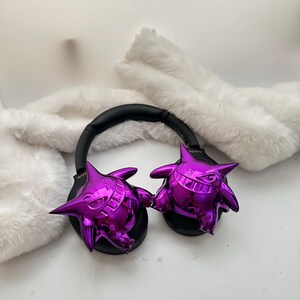 May include: Black headphones with a black headband and shiny purple Gengar figures on each ear cup. The headphones are resting on a white, fluffy surface.