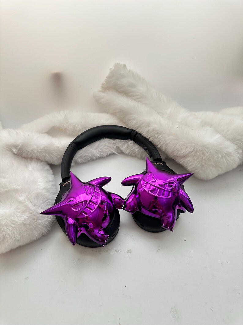 May include: Black headphones with a black headband and shiny purple Gengar figures on each ear cup. The headphones are resting on a white, fluffy surface.