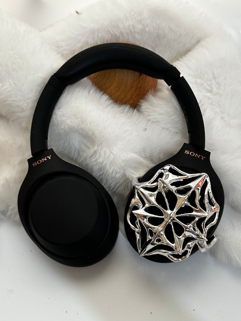 May include: Black Sony headphones with a silver decorative accent. The headphones are on a white, fluffy surface. The Sony logo is visible on the earcups.