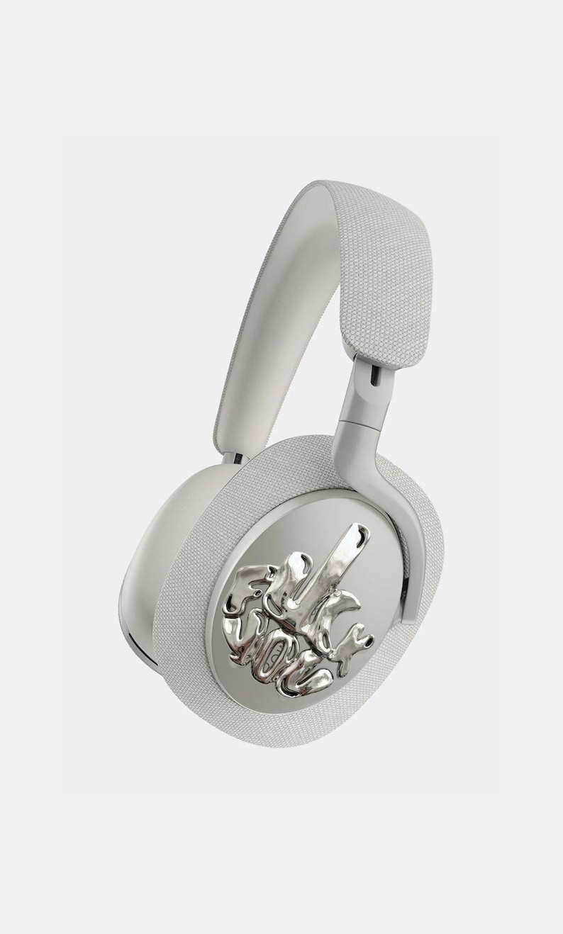 Puede incluir: Auriculares blancos de diadema con un emblema plateado en la orejera. El emblema presenta un dise&ntilde;o abstracto. La diadema y las almohadillas est&aacute;n acolchadas con una tela texturizada. Los auriculares est&aacute;n sobre un fondo blanco.