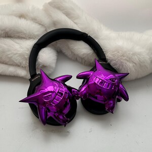 May include: Black Sony headphones with glossy purple Gengar figures attached to the earcups. The headphones rest on a white, fluffy surface. The Gengar figures have sharp teeth and pointed ears.