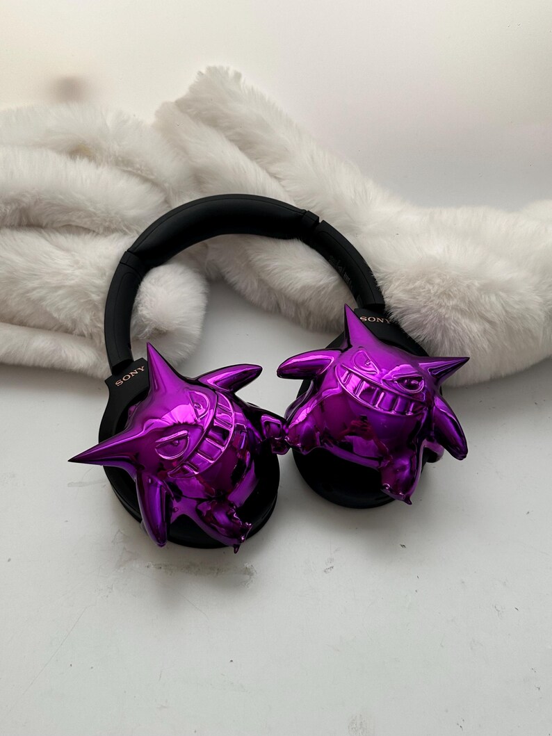 May include: Black Sony headphones with glossy purple Gengar figures attached to the earcups. The headphones rest on a white, fluffy surface. The Gengar figures have sharp teeth and pointed ears.