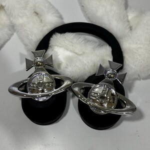 May include: A pair of black headband earmuffs with white faux fur ear covers. Each earmuff features a silver orb with a cross and a ring around it.